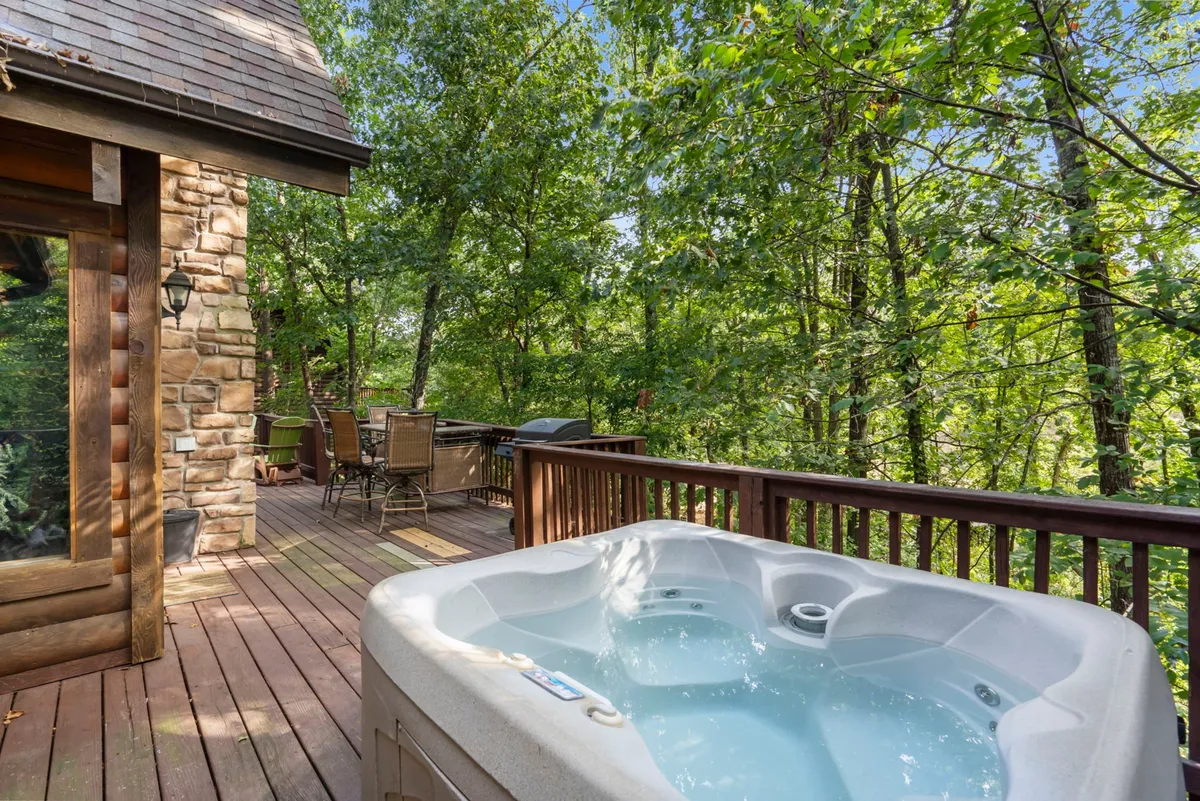 A tranquil wooden deck extends from the cabin, surrounded by lush green trees. The deck features a private hot tub set beneath the open sky, as well as a cozy outdoor seating area and a propane grill, all nestled amidst the wooded landscape.