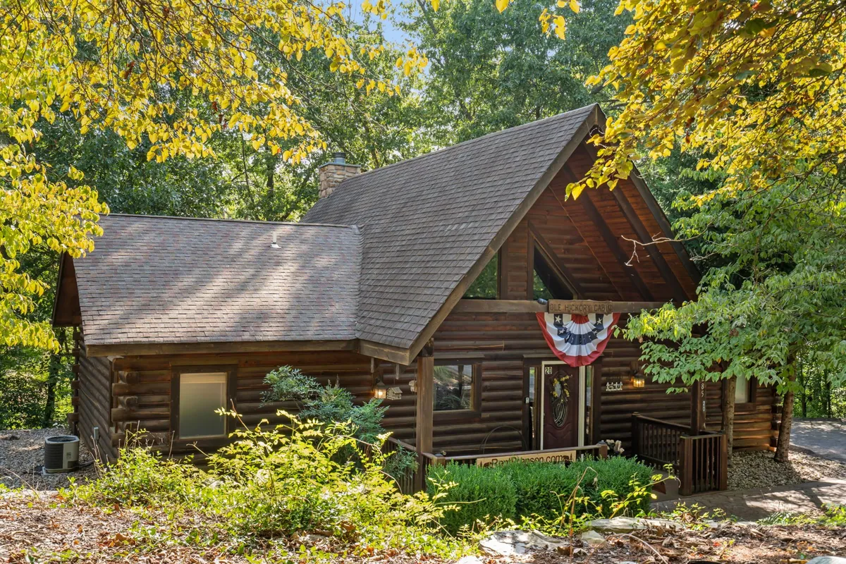 A cozy log cabin with green foliage and festive bunting on the porch creates a welcoming scene. The surrounding nature and inviting architecture evoke family gatherings and outdoor festivities, especially during seasonal events.