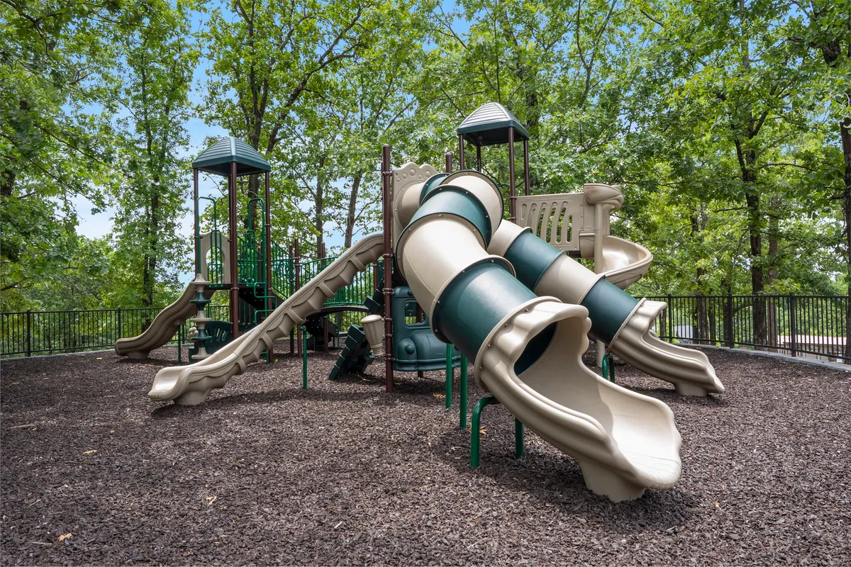 A colorful playground with slides and a climbing structure nestled among tall trees. The natural setting and cheerful equipment invite children to play and families to gather nearby.