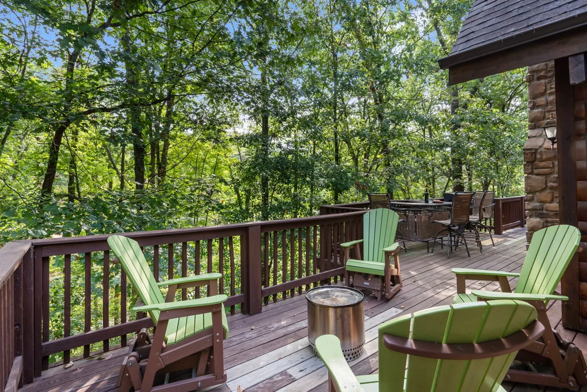 A charming deck with green Adirondack chairs is set against a backdrop of dense trees. The area includes a fire pit and is designed for socializing, relaxing, and enjoying the outdoors at any time of day.