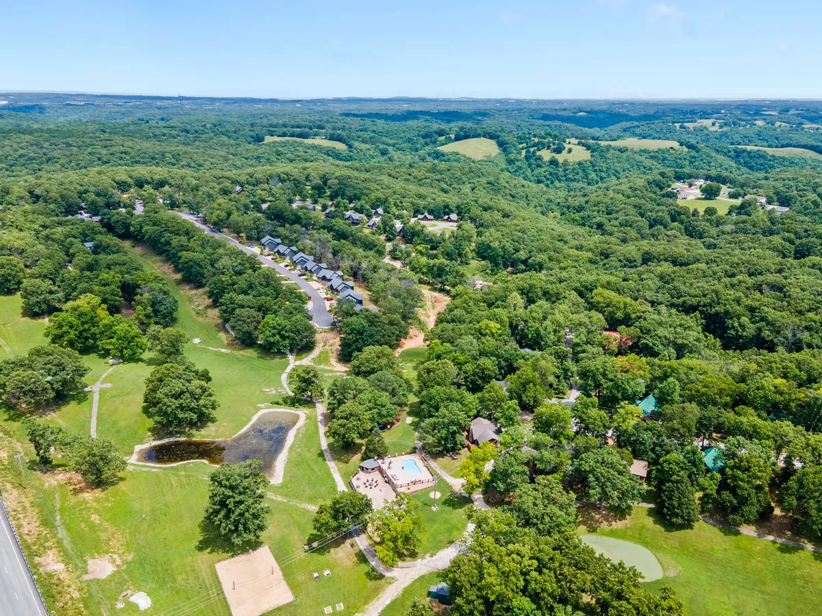 An exterior, wide-angle view of spacious green lawns, clusters of trees, a small pond, and a community pool area, with lodges along a winding pathway. The vibrant setting highlights both natural and communal amenities.