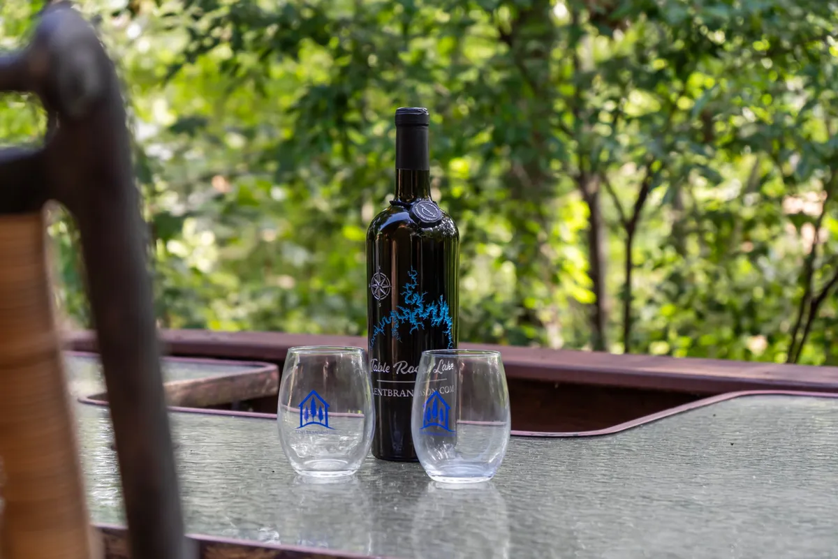 A close-up of a bottle of wine and two glasses set outdoors, suggesting an intimate moment for two. The setting is warm and inviting, with an atmosphere suited for relaxation and connection under open skies.