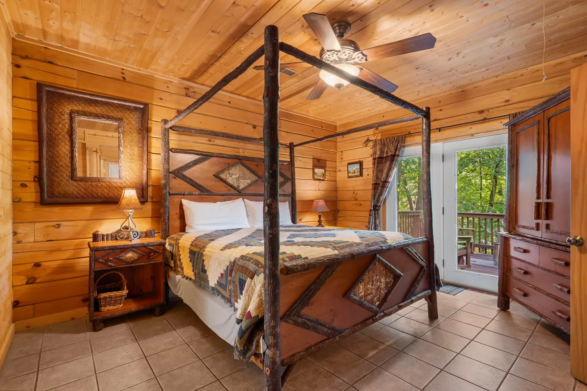 A beautifully appointed bedroom featuring a four-poster king-sized bed, warm wooden furniture, and a large window that provides a peaceful view of the surrounding woods. Sunlight pours into the room, highlighting the inviting bedding and creating a cozy, restful ambiance.