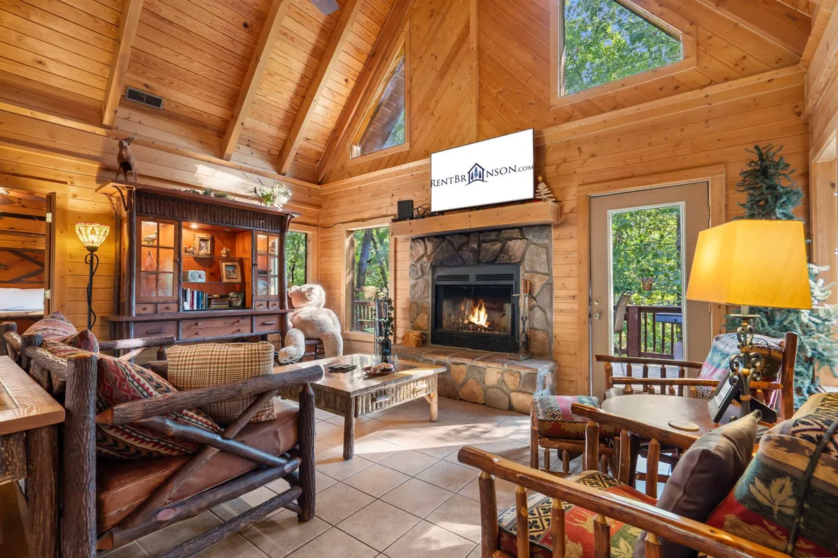 The image showcases a warm, inviting cabin living area featuring a crackling fireplace, rustic wooden furniture, expansive windows framing wooded views, and a stylish entertainment center. Sunlight pours in, creating a cozy atmosphere perfect for relaxing, reading, or gathering with friends and family.