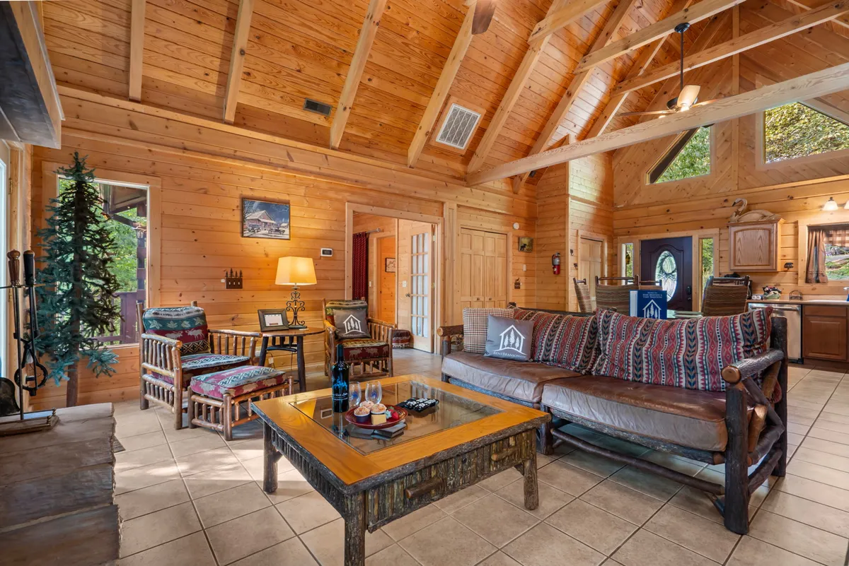 A spacious great room featuring exposed wooden ceiling beams, a cozy sofa with patterned cushions, a rustic coffee table, and a decorative lamp. Large windows fill the room with natural light, and the warm wood tones create a welcoming, homey atmosphere.