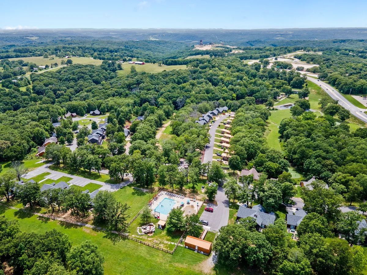 This image captures an inviting swimming pool surrounded by lounge chairs, open fields, and clusters of cabins nestled among the trees. The relaxed atmosphere and communal layout highlight the resort environment and amenities available to families.