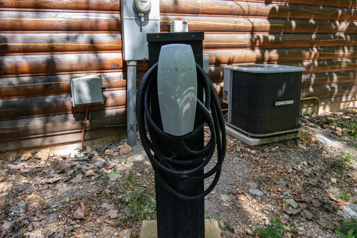 A close-up of a Tesla charging station installed next to a log cabin exterior, surrounded by greenery. The image blends rustic cabin architecture with modern technology, emphasizing eco-friendly convenience.