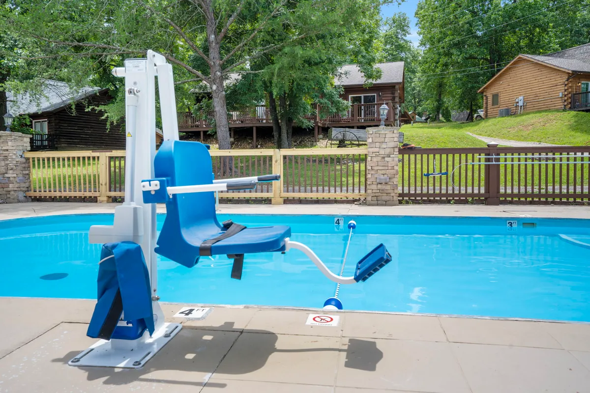 A sunlit swimming pool area surrounded by greenery, featuring a pool lift for accessibility and wooden cabins visible in the background. The scene emphasizes both relaxation and inclusivity, with a clear view of the resort’s natural surroundings despite nearby construction.