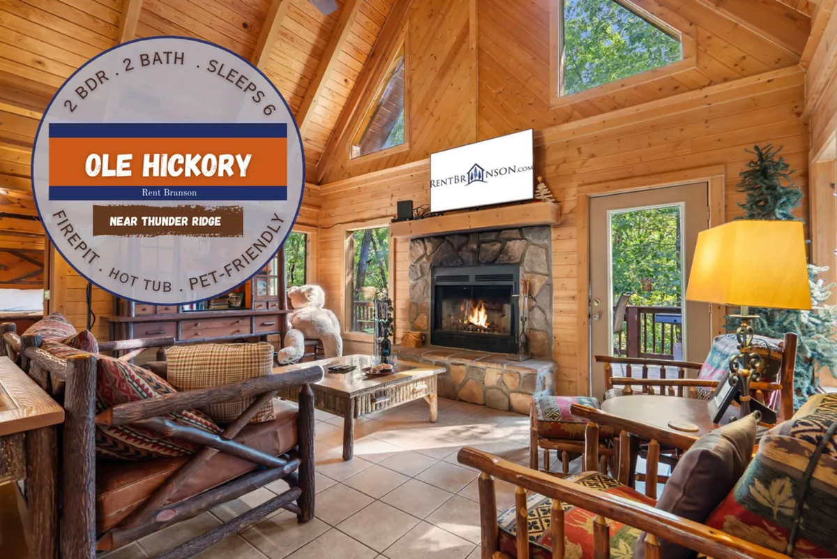 A warm, inviting living space with a prominent stone fireplace, large windows looking out to nature, wooden ceiling beams, and comfortable seating arranged for conversation or relaxation.